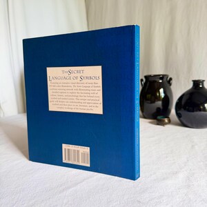 May include: A hardback book titled "The Secret Language of Symbols" with a blue cover. The book's title and description are printed in white. Two dark vases are in the background.