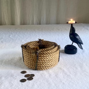 May include: A woven rope basket and a black bird-shaped candle holder with a lit candle. Several small, round, bronze-colored coins are scattered near the basket. The items are arranged on a white surface.