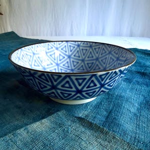 May include: A ceramic bowl featuring a repeating blue and white geometric triangle pattern across its interior and exterior surfaces. The bowl has a smooth, rounded shape with a dark rim and a solid white base, showcasing a clean, symmetrical design.