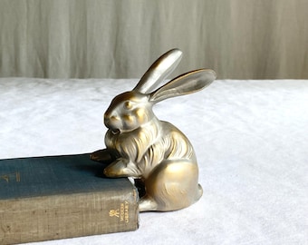 May include: A gold and silver-toned rabbit figurine, posed in a seated position, with long ears and detailed fur. The rabbit is positioned next to a vintage blue book. The figurine is a decorative item.