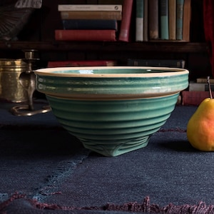 May include: A teal ceramic bowl with a ribbed exterior and a wide, flat rim. The bowl features a smooth, glossy glaze and a tapered base. A single yellow and red pear sits to the right of the bowl on a dark blue textured fabric surface.