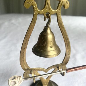 May include: A brass bell with a decorative stand. The bell hangs from a lyre-shaped frame. A small skull-shaped striker is included. The bell and stand have an aged, antique appearance.