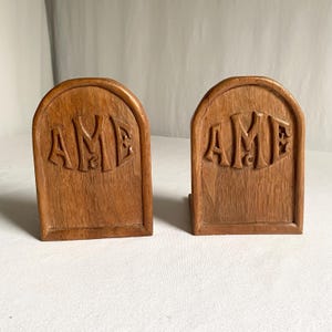 May include: Two wooden bookends with a light brown finish. Each bookend has an arched top and the letters "AME" carved into the front. The bookends are designed to hold books upright on a shelf.