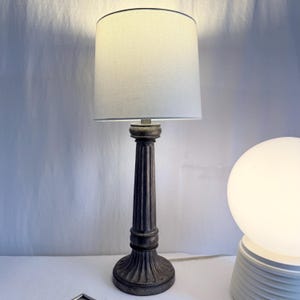 May include: A decorative table lamp with a cylindrical lampshade and a dark, fluted column base. The lampshade is a light cream color, and the base has an antique finish. The lamp is designed for indoor use and provides ambient lighting.