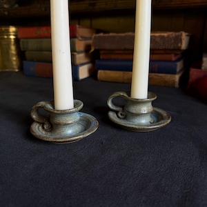May include: Two ceramic candle holders with a speckled blue and brown glaze, each featuring a small side handle and a circular base. Each holder contains a tall, slender, off-white taper candle standing upright in the center.