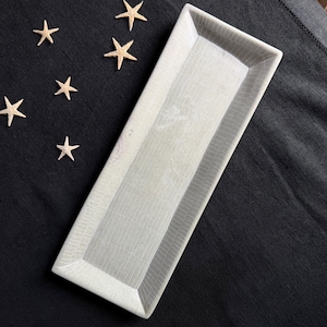 May include: A rectangular ceramic tray with a light gray, crackled glaze finish and a textured surface pattern. The tray is positioned diagonally alongside several small, light-colored starfish arranged on a dark, solid-colored surface.