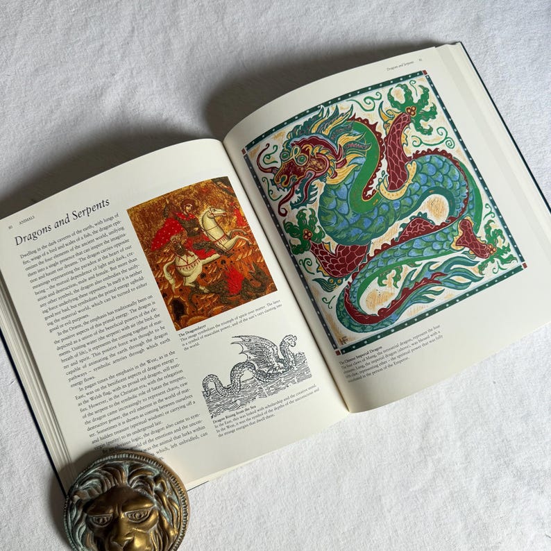 May include: An open book displaying a colourful illustration of a dragon in green, red, and blue. The left page has text titled "Dragons and Serpents" and an image of a person on a horse. A brass lion head is visible.