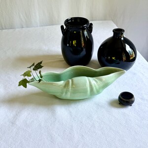 May include: A collection of ceramic vases and a decorative bowl on a white surface. The vases are black and glossy, with one having handles. A light green, leaf-shaped bowl holds greenery. A small, dark bowl sits nearby.