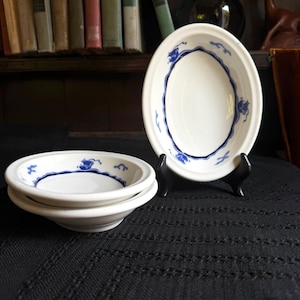 May include: Three white ceramic bowls with a smooth finish and a blue painted decorative border featuring small abstract shapes. One bowl is displayed upright on a black stand, while two other bowls are stacked horizontally in the foreground.