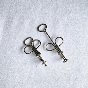 May include: Two silver-toned antique medical tools, possibly surgical instruments, are displayed against a white background. Each tool has a handle, a central body, and a plunger-like end. The tools are likely used for medical procedures.