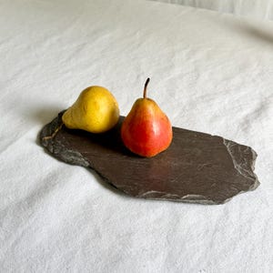 May include: A rectangular slate serving board with a rough, natural edge. Two pears are placed on top of the dark grey stone surface: one is a solid yellow pear lying on its side, and the other is an upright pear with a gradient of red and yellow.