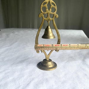 May include: A vintage brass bell with a decorative stand. The bell is suspended within a curved frame, and the stand has a round base. The bell is approximately 2 inches tall, and the stand is about 6 inches tall.