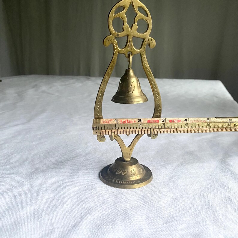May include: A vintage brass bell with a decorative stand. The bell is suspended within a curved frame, and the stand has a round base. The bell is approximately 2 inches tall, and the stand is about 6 inches tall.