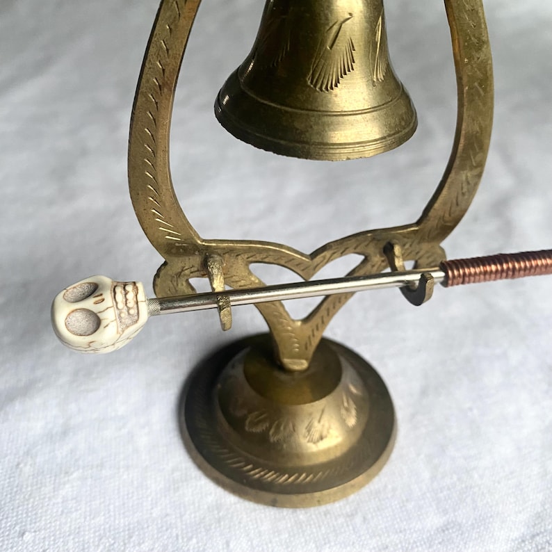 May include: A vintage brass bell with a skull-shaped clapper. The bell is mounted on a decorative stand with a heart-shaped design. The clapper handle is wrapped with copper wire.