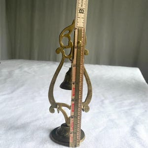 May include: A vintage brass bell with an ornate, openwork frame. The bell is approximately 7 inches tall, measured with a wooden ruler. The base is round with decorative detailing, and the bell itself is dark.