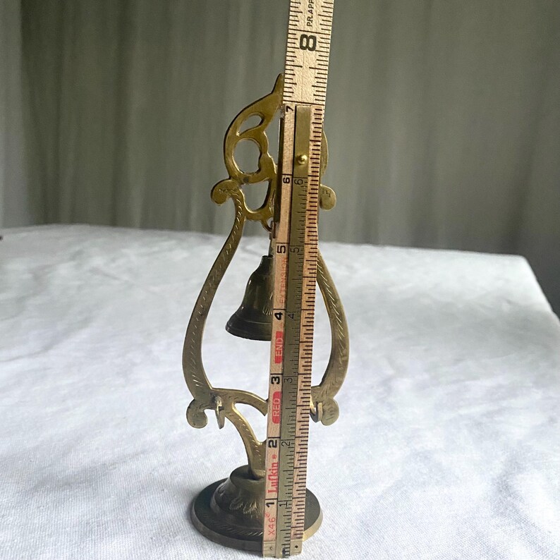 May include: A vintage brass bell with an ornate, openwork frame. The bell is approximately 7 inches tall, measured with a wooden ruler. The base is round with decorative detailing, and the bell itself is dark.