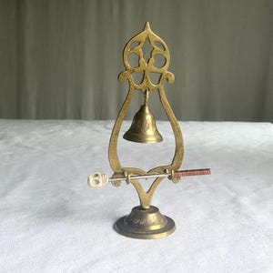 May include: A vintage brass bell with a decorative frame and a small clapper. The bell is suspended within an ornate, openwork structure. A small, decorative handle is attached to the bell's base. The bell is likely used for decorative purposes.