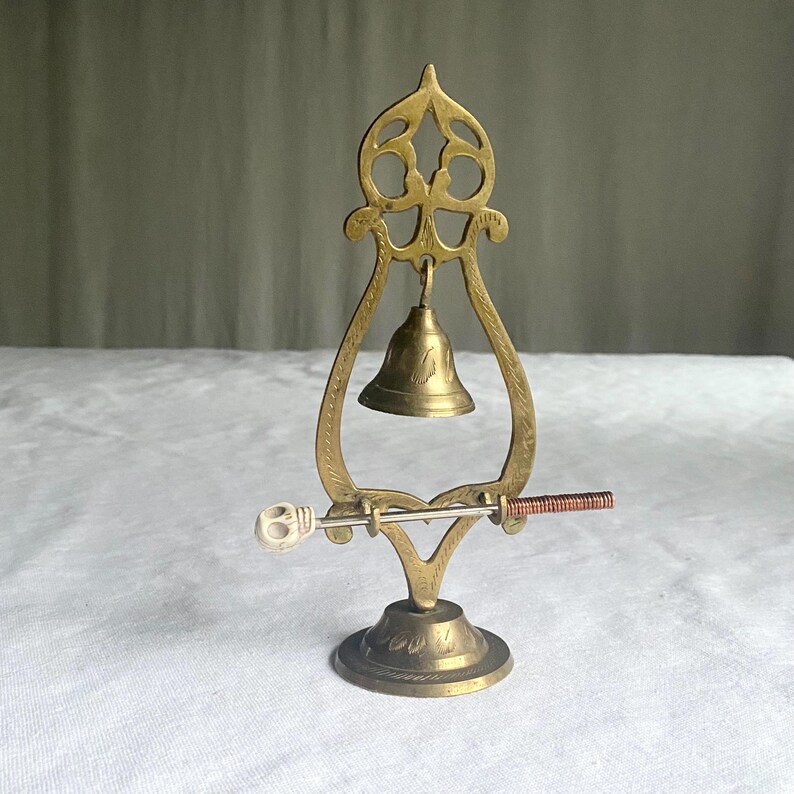 May include: A vintage brass bell with a decorative frame and a small clapper. The bell is suspended within an ornate, openwork structure. A small, decorative handle is attached to the bell's base. The bell is likely used for decorative purposes.
