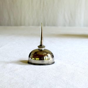 May include: A small, vintage silver oil can with a rounded body and a long, pointed spout. The can has a reflective surface, and the spout is a lighter silver colour. The oil can is likely used for lubricating machinery or other mechanical devices.