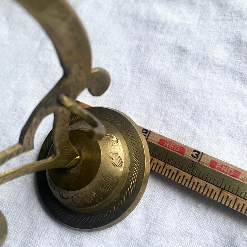 May include: Close-up of a brass decorative object with a circular base and an ornate design. A wooden ruler with red text is next to the object. The ruler measures in inches.