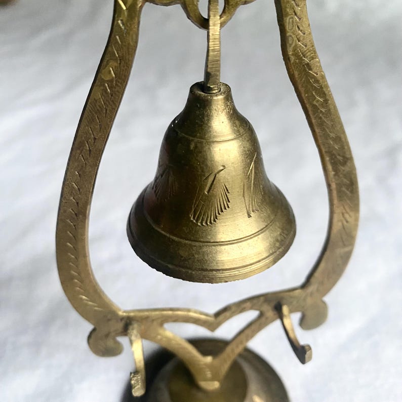 May include: A small, antique brass bell with a decorative etched design, suspended within an ornate brass frame. The bell has a classic shape and is suspended from a curved, heart-shaped support. The frame features etched patterns.