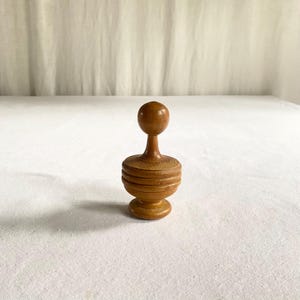 May include: A small, turned wooden object with a rounded top, a slender stem, and a ribbed, cylindrical base. The wood has a warm, medium brown tone. The object is likely a decorative item or a small kitchen tool.