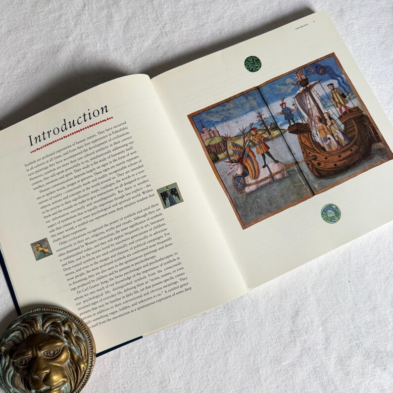 May include: An open book displaying the title "Introduction" on the left page. The right page shows a vibrant illustration of a ship and figures. The book rests on a white surface, with a decorative lion head in the lower left corner.