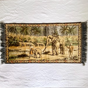 May include: Rectangular tapestry featuring a detailed scene with elephants, palm trees, and figures in a tropical landscape. The tapestry has a dark brown border with decorative patterns and fringe on the short ends. The color palette includes beige, brown, and green.