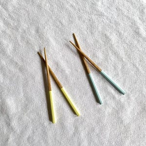 Könnte beinhalten: Vier Paar Essstäbchen aus Holz mit lackierten Spitzen. Zwei Paare haben gelbe Spitzen und zwei hellblaue Spitzen. Die Essstäbchen sind auf einer weißen, strukturierten Oberfläche angeordnet.