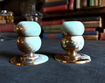 May include: Two matching candle holders featuring a polished gold-tone metal base and stem, topped with a rounded, matte light blue ceramic element. The metal surfaces show a weathered, oxidized patina with visible green and white textured spots.