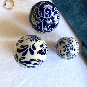 Puede incluir: Tres esferas decorativas de cerámica con patrones florales y abstractos azules y blancos. Las esferas varían en tamaño y diseño, con detalles intrincados. La esfera más grande presenta un complejo patrón de remolino azul y blanco.