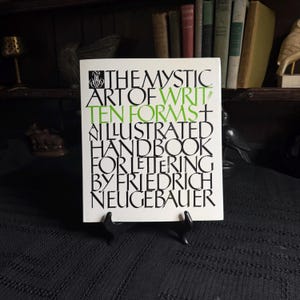 May include: A white book cover titled The Mystic Art of Written Forms, an illustrated handbook for lettering by Friedrich Neugebauer. The text is printed in black with the word written in green, featuring a decorative emblem at the top left.