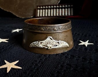 May include: A circular bronze bowl featuring a raised white relief of a bat with outstretched wings on the front. The upper rim is decorated with an embossed vine and leaf pattern. The metal surface has a dark, aged patina throughout.