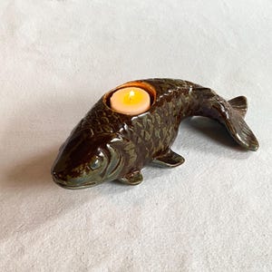 May include: A ceramic fish-shaped candle holder in brown and green hues. The candle holder has a textured surface and a cavity for a lit candle. The fish's tail is curved, and the candle emits a warm glow.