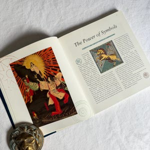 May include: An open book displaying the title "The Power of Symbols". The left page shows a vibrant illustration of figures in traditional attire. A brass lion-head bookend is visible at the bottom.