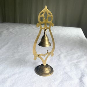 May include: A small, ornate brass bell suspended within a decorative brass frame. The frame features an openwork design at the top and a floral pattern on the base. The bell is suspended from a hook and has a simple, classic shape.