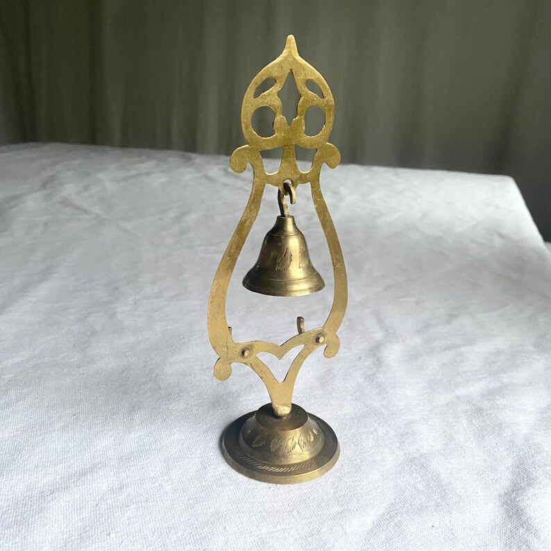 May include: A small, ornate brass bell suspended within a decorative brass frame. The frame features an openwork design at the top and a floral pattern on the base. The bell is suspended from a hook and has a simple, classic shape.