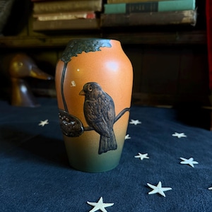 May include: A ceramic vase with a gradient finish transitioning from orange at the top to dark green at the base. The vase features a raised, dark brown relief of a bird perched on a branch with a textured, dark brown leafy element above it.