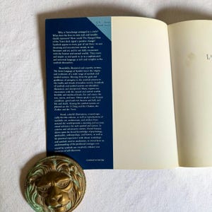 May include: A book with a blue cover and a bronze lion medallion. The book's cover has text about symbols and the Tarot. The medallion is round with a detailed lion's face. The book is open to a blank page.