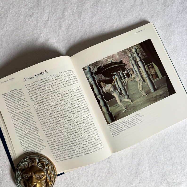 May include: An open book titled "Dream Symbols" with a surrealist painting on the right page. The painting depicts figures in a dreamscape. A brass lion-head bookend is at the bottom of the book.
