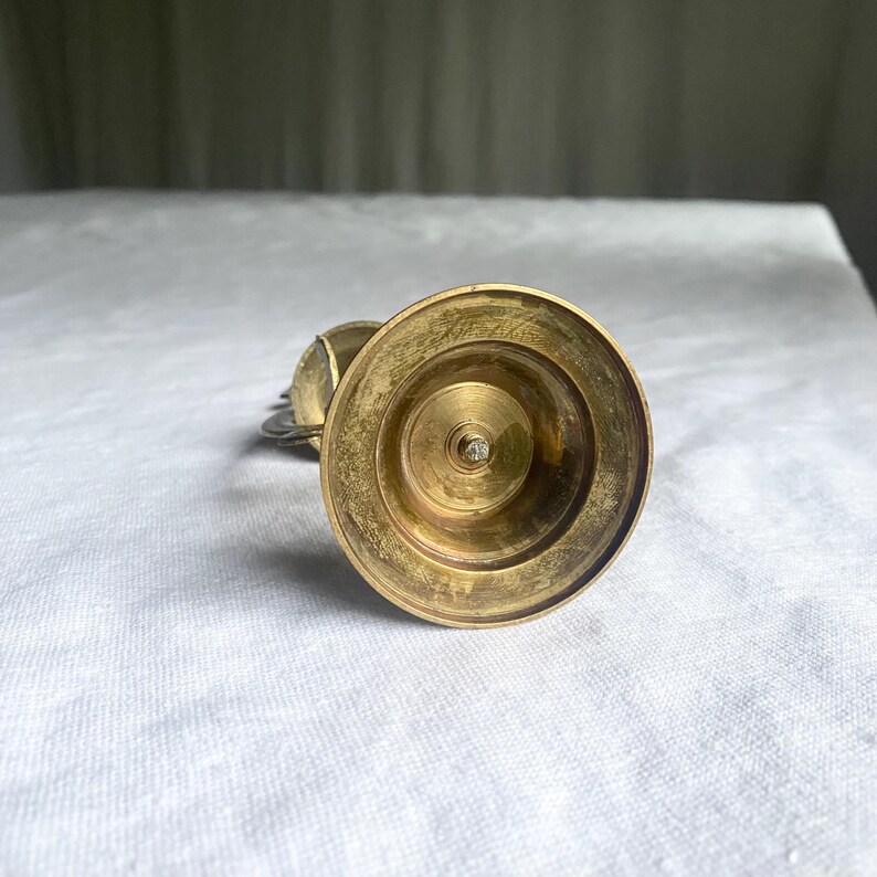 May include: A vintage brass candlestick holder with a wide, circular base and a central spike. The candlestick has a warm, golden hue and shows signs of age. The design is simple and elegant, suitable for various decor styles.