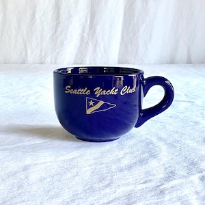 May include: A deep blue ceramic mug with a rounded shape and a handle. The mug features the text "Seattle Yacht Club" in gold script, along with a gold emblem of a flag with a star. The mug is set against a white background.