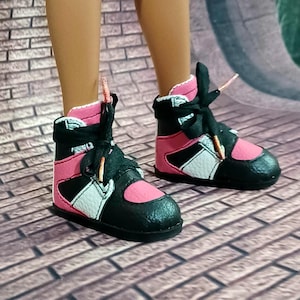 May include: A pair of miniature high-top sneakers in black, pink, and white. The shoes have black laces and a pink tongue, with a white accent on the side. The soles are black, and the shoes appear to be made of leather or a similar material.