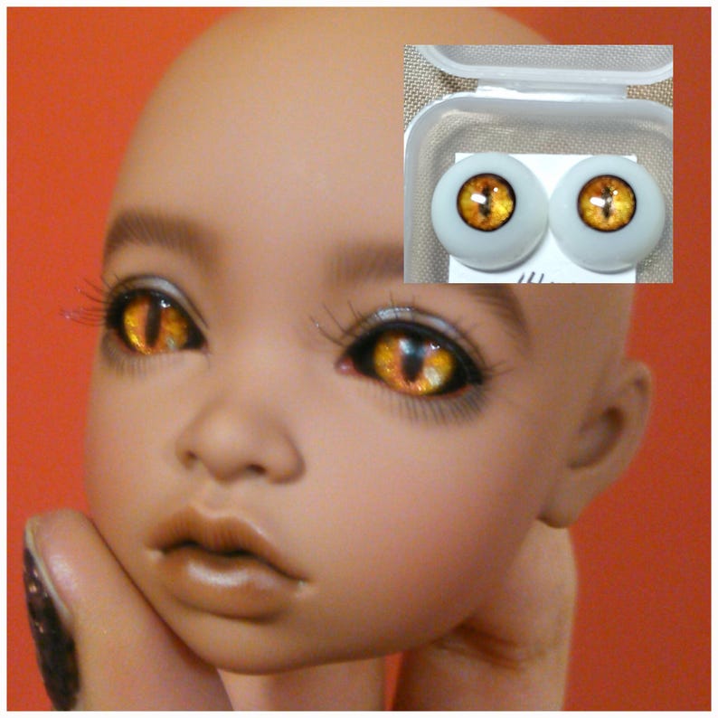 Puede incluir: Un par de ojos de mu&ntilde;eca naranja y amarillo con un dise&ntilde;o de ojo de gato. Los ojos est&aacute;n en un contenedor de pl&aacute;stico transparente con una tapa blanca.
