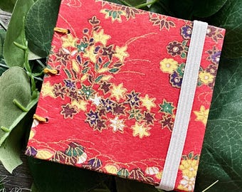 May include: A small, square red notebook with a floral design in yellow, green, and white. The notebook has a white elastic band closure and gold binding. The notebook is surrounded by green leaves and white fluffy grass.