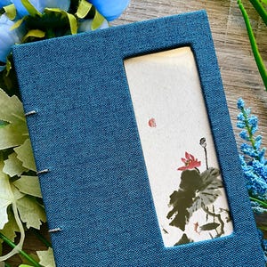 May include: A blue fabric-covered journal with a rectangular window revealing a watercolor painting of a lotus flower. The journal is bound with silver staples. The painting features red, green, and black colors.