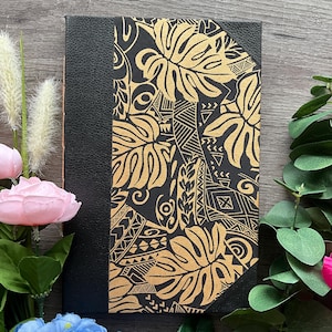 May include: A black journal featuring a gold leaf pattern on the cover. The journal has a leather spine and a decorative floral design. The cover is adorned with a repeating pattern of stylized leaves and geometric shapes.