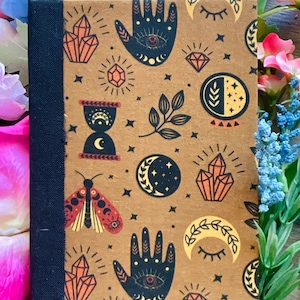 May include: A hardback journal with a brown cover featuring celestial and mystical illustrations. The cover design includes hands with eyes, crystals, moons, and a moth. The spine is a dark blue fabric.