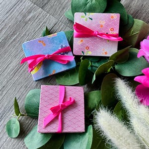 May include: Three small, square gift boxes with pink ribbons, each featuring a different Japanese-inspired pattern. The boxes are surrounded by greenery and pink flowers, creating a decorative presentation.
