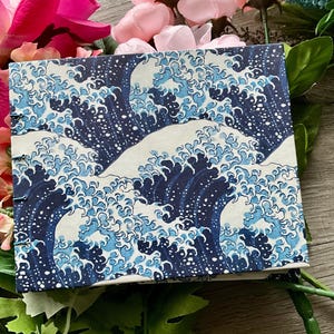 May include: A square, bound notebook featuring a repeating pattern of stylized ocean waves in shades of blue and white. The notebook's cover design is reminiscent of traditional Japanese art. The edges are visible, showing the pages are bound together.